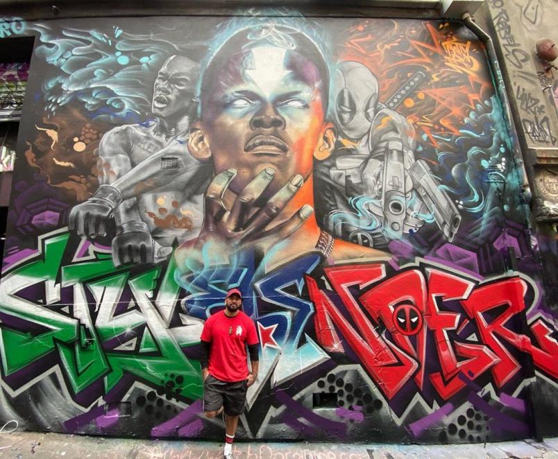 The Artist Poses In Front Of His Adesanya Mural. From His Instagram @mattybro_art