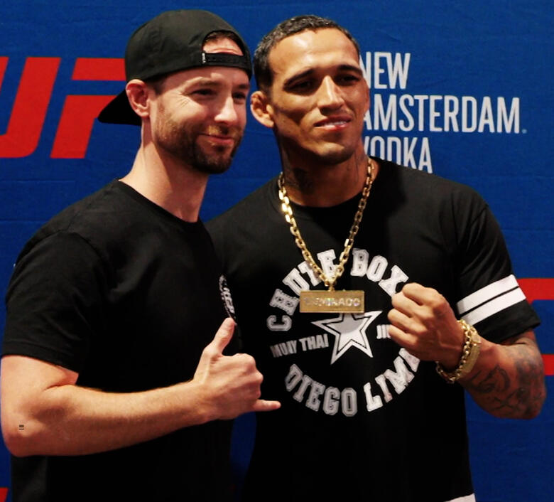 UFC fighter Charles Oliveira poses with a fan at a meet-and-greet event. Oliveira, wearing a black ‘Chute Boxe Diego Lima’ t-shirt and a gold chain with a ‘Champion’ pendant, smiles and makes a fist gesture. The fan, dressed in a black t-shirt and a backward black cap, holds up a shaka sign. A blue UFC-branded backdrop with sponsor logos is visible behind them, and another attendee is capturing the moment on a smartphone.