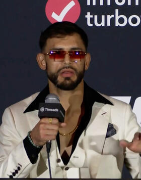 UFC fighter Yair Rodriguez speaks at the UFC 314 post-fight press conference. He is wearing a white suit with a black shirt and gold chain, accessorized with red-tinted sunglasses. Rodriguez holds a microphone labeled “Threads” as he addresses the media in front of a backdrop featuring UFC 314 branding, sponsor TurboTax, and the fight card listing Volkanovski vs. Lopes. A can of DRIP energy drink is visible on the table in front of him.
