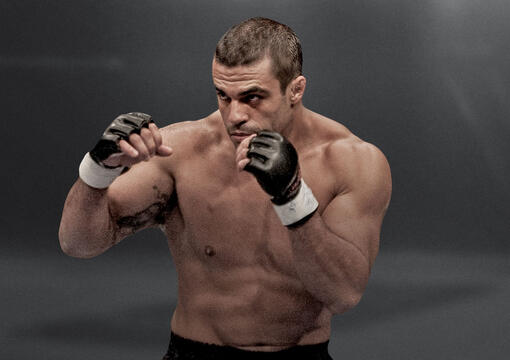 “Vitor Belfort, shirtless and wearing MMA gloves, stands in a fighting stance with his fists raised, ready for action against a dark, dramatic background.